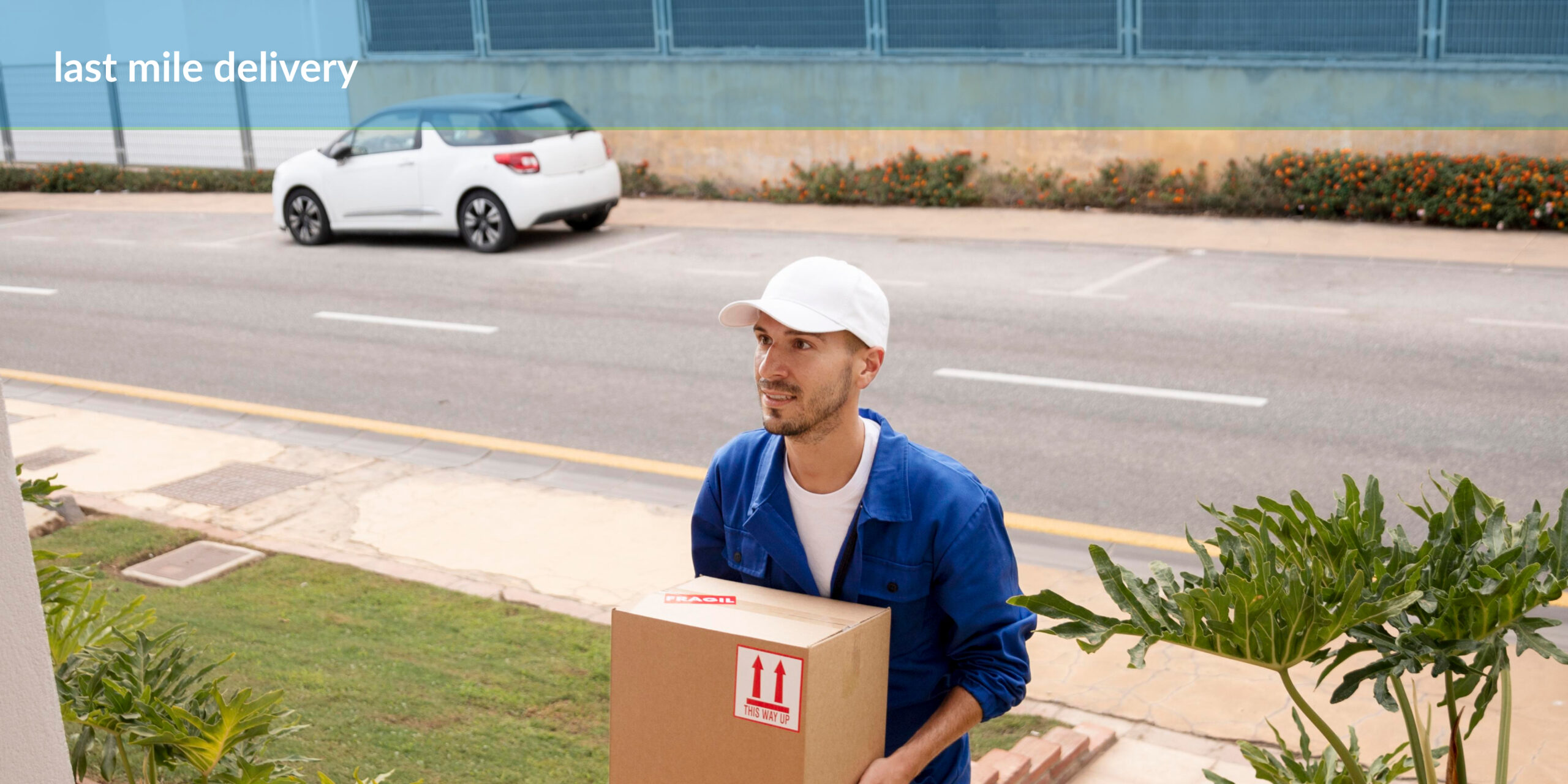 A delivery person in a blue uniform and white cap holding a cardboard box with a "This way up" sticker standing on the sidewalk next to a road with a white car passing by, with text "last mile delivery" in the upper left corner. Greenery is visible in the foreground and background.