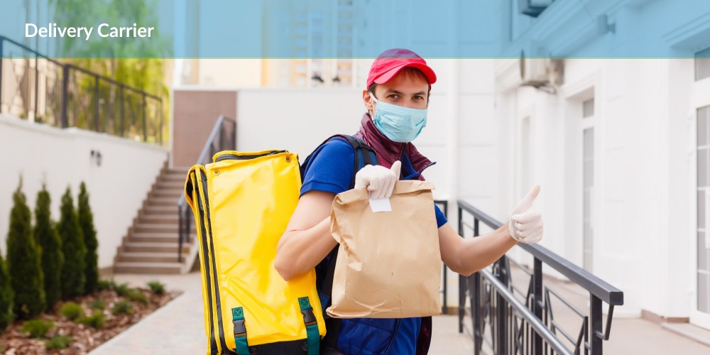 Delivery carrier : A person in a red cap and blue shirt, wearing a mask and gloves, holding a paper bag and giving a thumbs-up outside a building.