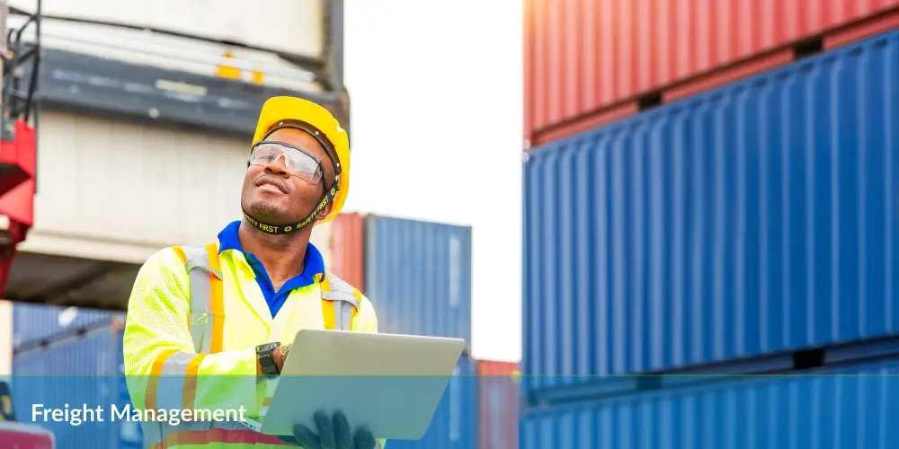 Worker in hi-vis gear using laptop at a shipping container yard.