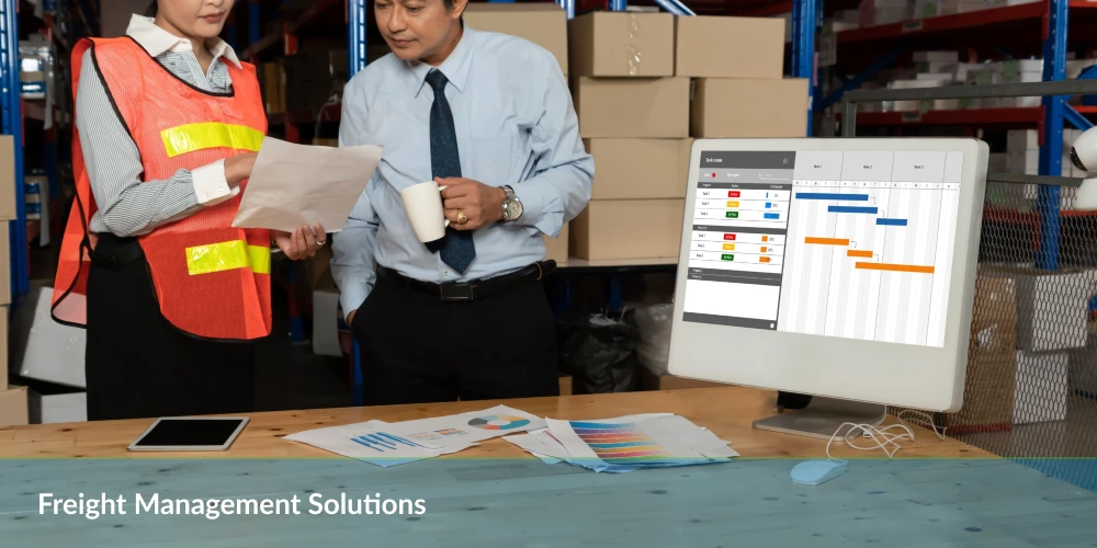 Freight Management Solutions - Two workers with documents in a warehouse and a computer screen displaying a schedule.