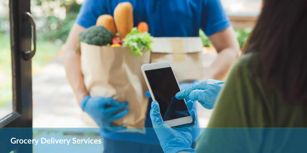 Delivery person in blue gloves handing over groceries to customer checking phone at doorway.