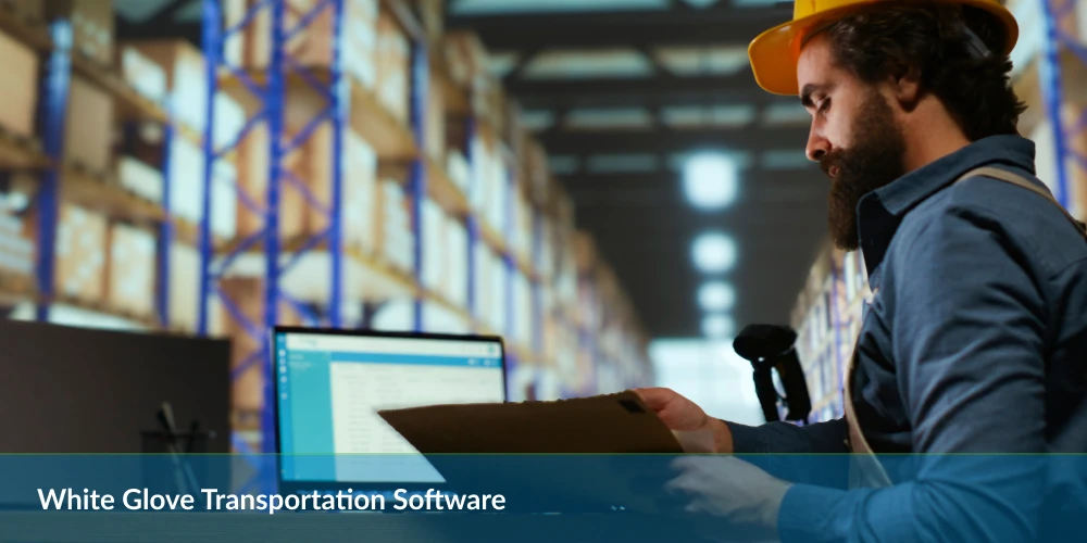 Worker in hard hat checking inventory in a warehouse with transportation software on screen.