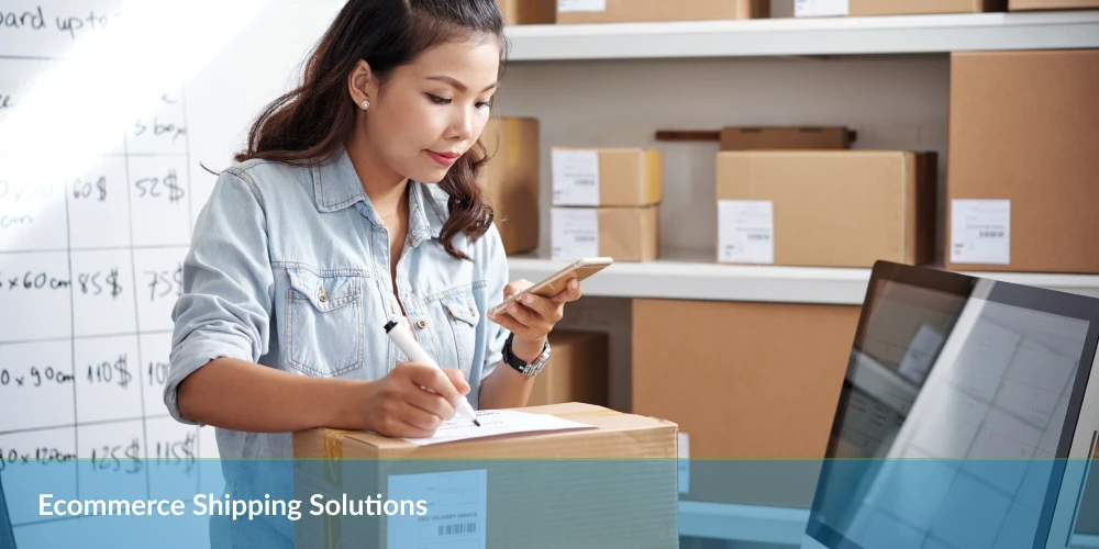A person writes on a cardboard box while checking their phone, surrounded by shipping boxes and a computer in a warehouse setting.